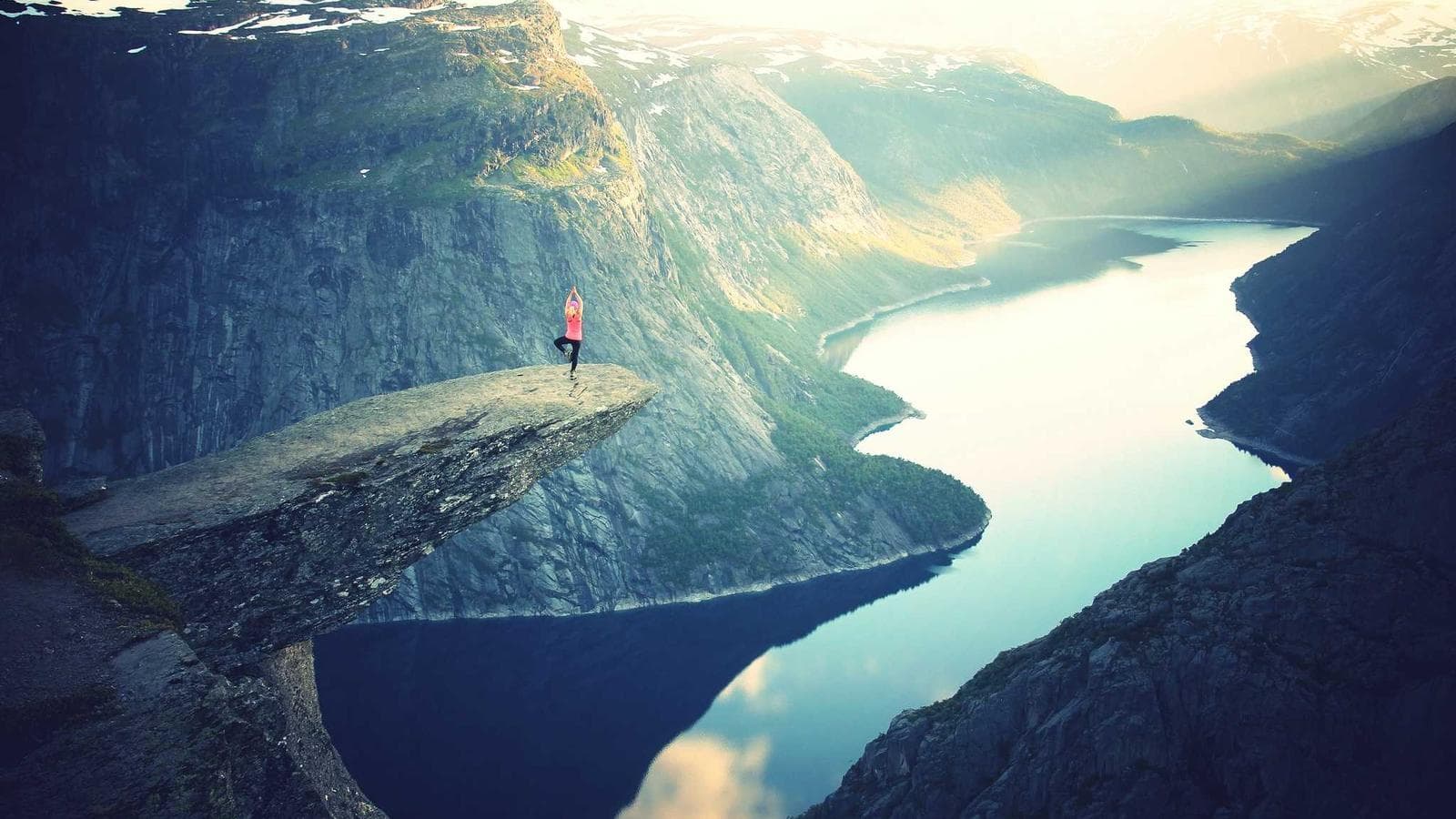 A person in a pink shirt and dark pants stands in a yoga pose on a precarious rock formation overlooking a vast lake and mountainous landscape.