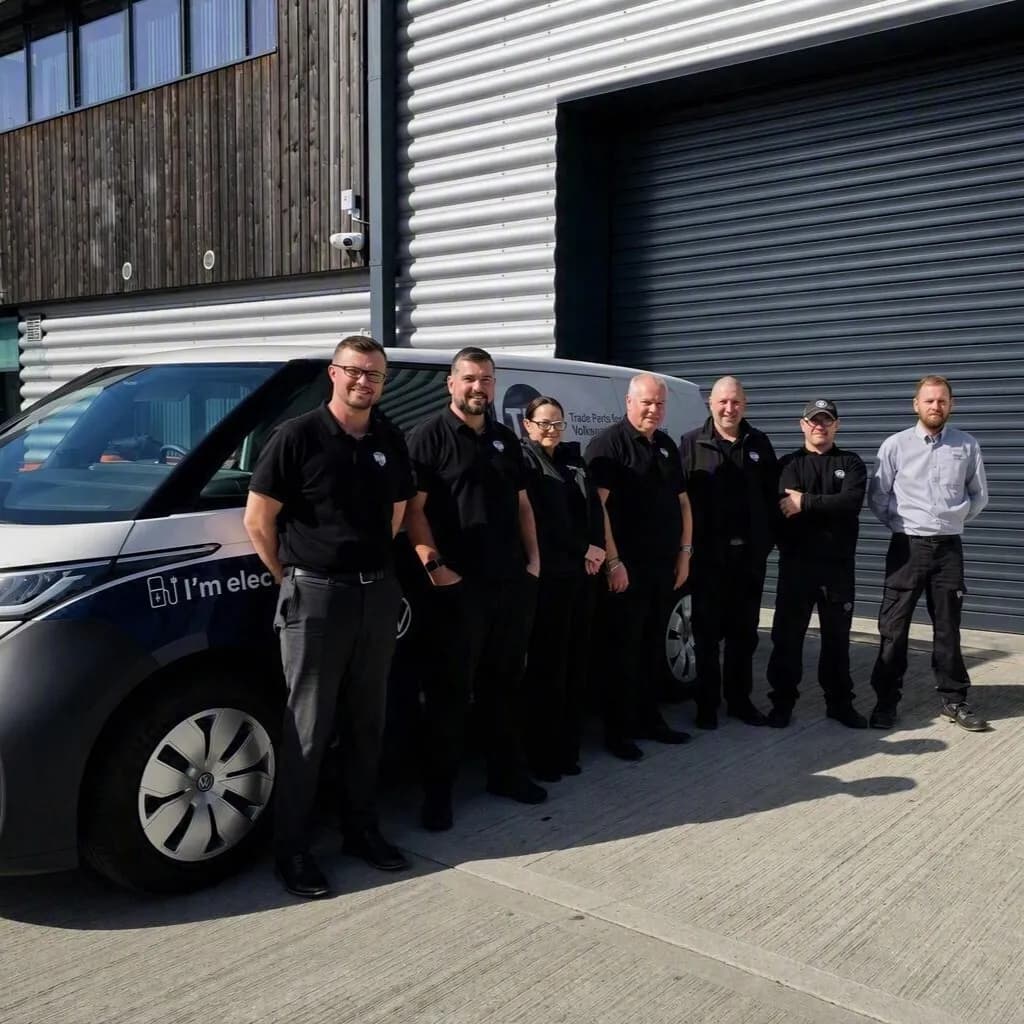 Seven people in uniforms standing beside a white van with text on it, in front of a building with a metal facade and a garage door.
